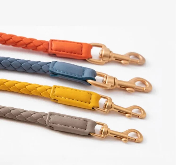 Four colorful braided leather straps in orange, slate blue, mustard yellow, and charcoal grey, each featuring a matte gold-toned swivel lobster clasp, displayed against a clean white background.