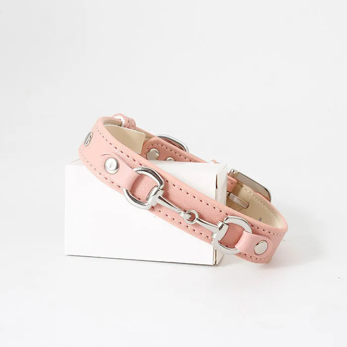 A light pink leather pet collar featuring decorative silver horse-bit hardware and round metal studs, presented on a small white box against a plain background.